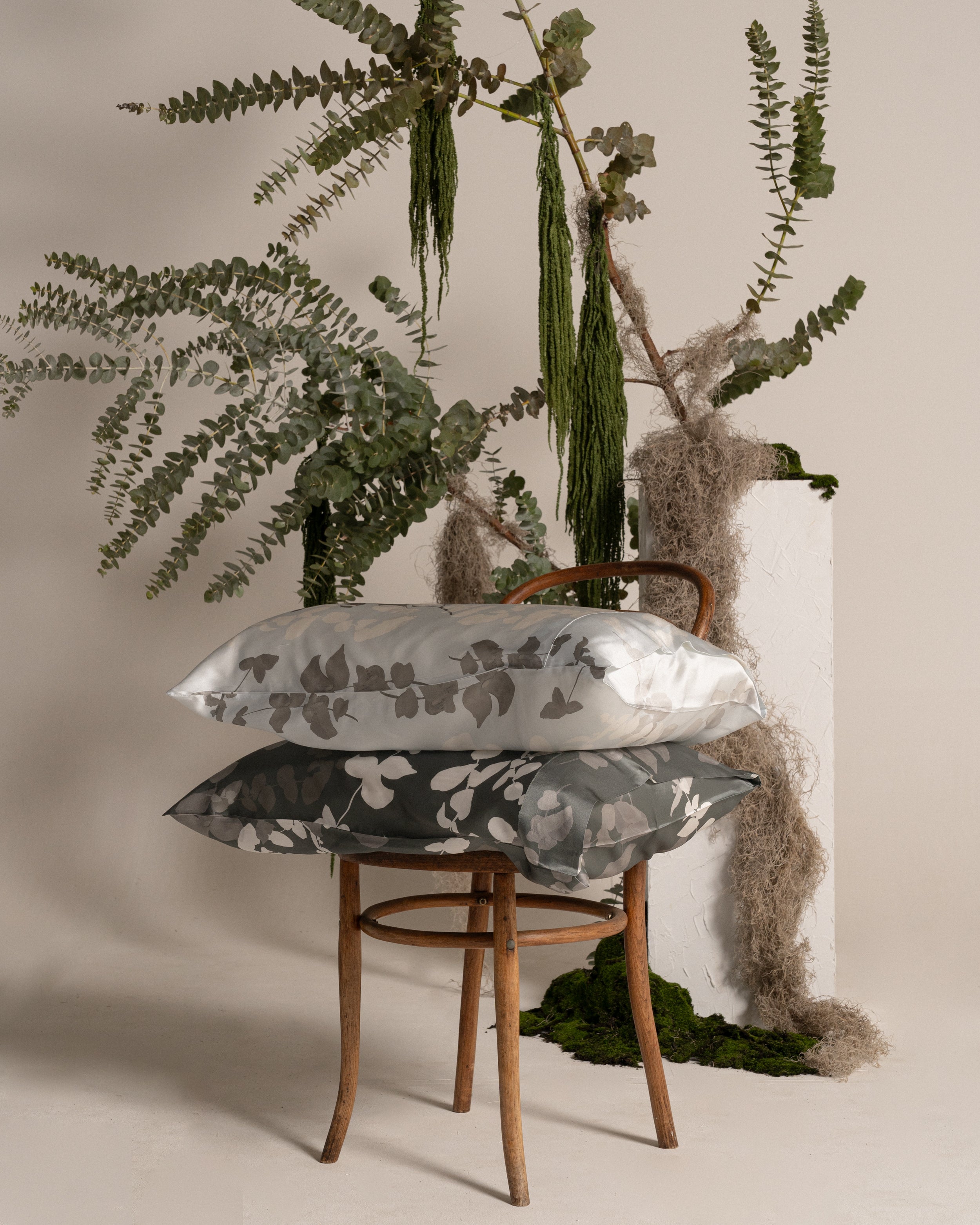 Two silk pillows on a wooden chair with plants in the background.